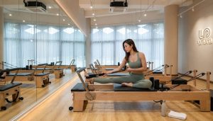 Woman performing a seated Pilates reformer exercise in a bright studio with mirrored walls, wooden reformer machines, sheer curtains, and LA Pilates branding on the wall.