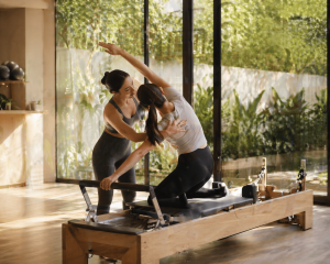 Pilates instructor assisting a client with a side stretch on a reformer machine in a sunlit studio.
